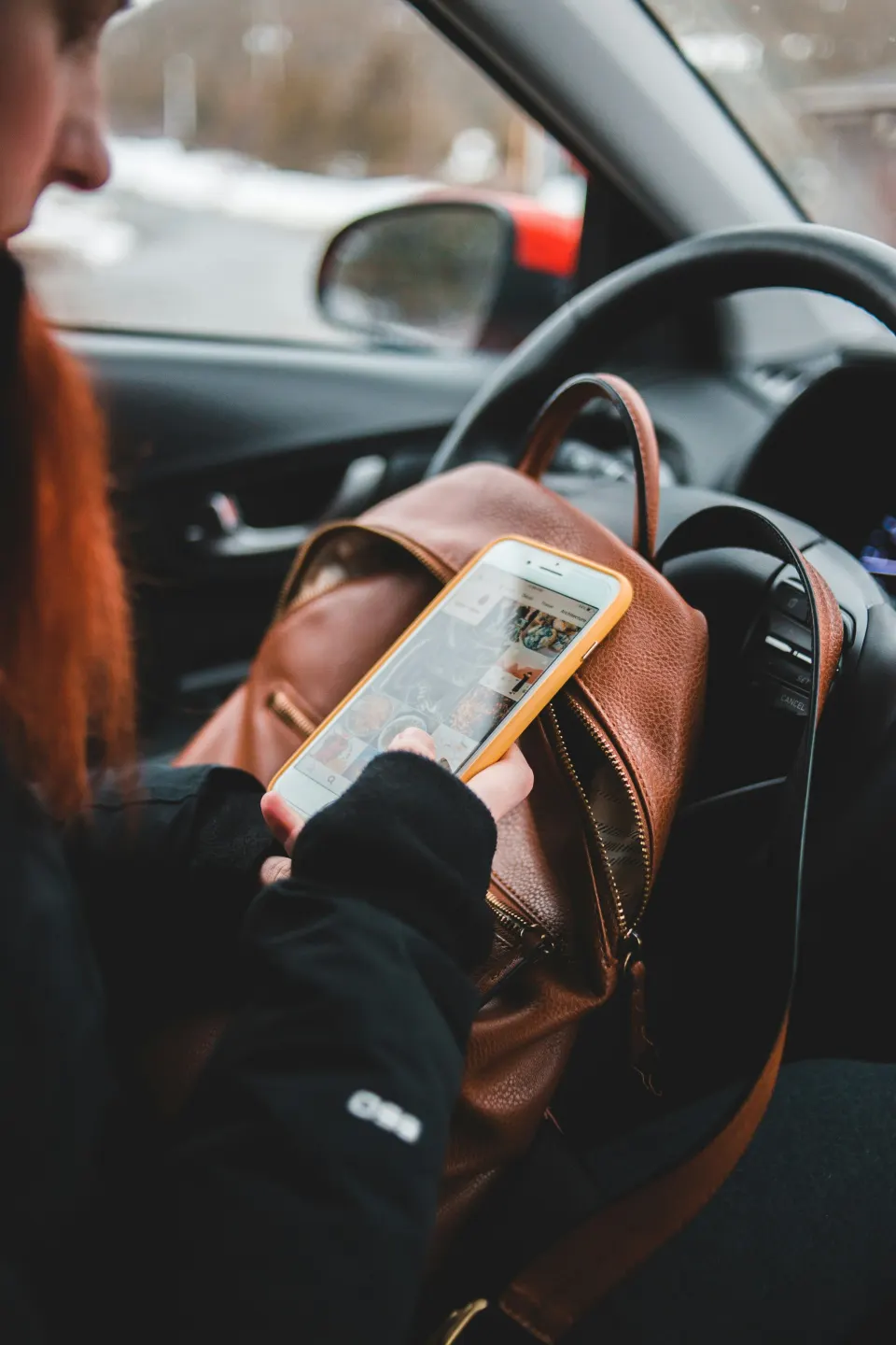 Person sitting in a car looking at a smartphone with a gallery of photos, with a brown leather bag on their lap.