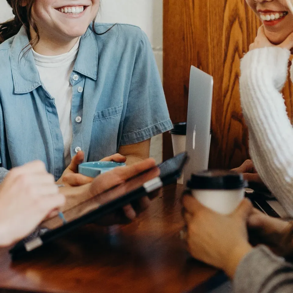 Two women sitting at a table with laptops and coffee cups, smiling and talking.