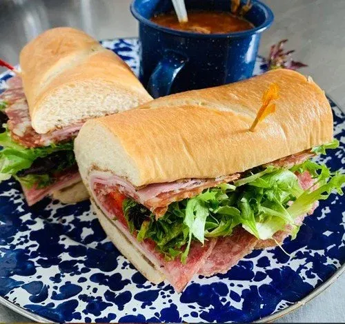 A plate with two turkey and ham sandwiches on hoagie rolls with lettuce and cold cuts, accompanied by a blue mug of soup or chili.